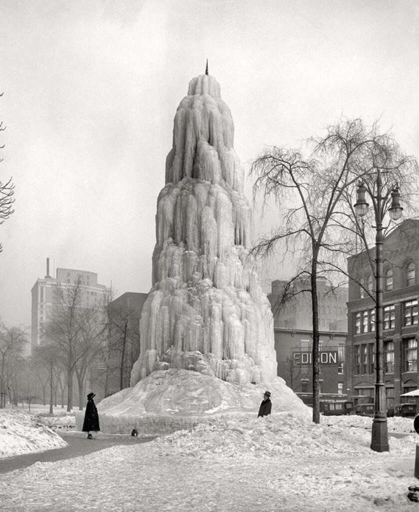 A towering ice sculpture in a park, resembling a frozen fountain, stands amidst snow. Two people in long coats are nearby, one with a dog. Leafless trees and buildings surround the scene, creating a wintry atmosphere.