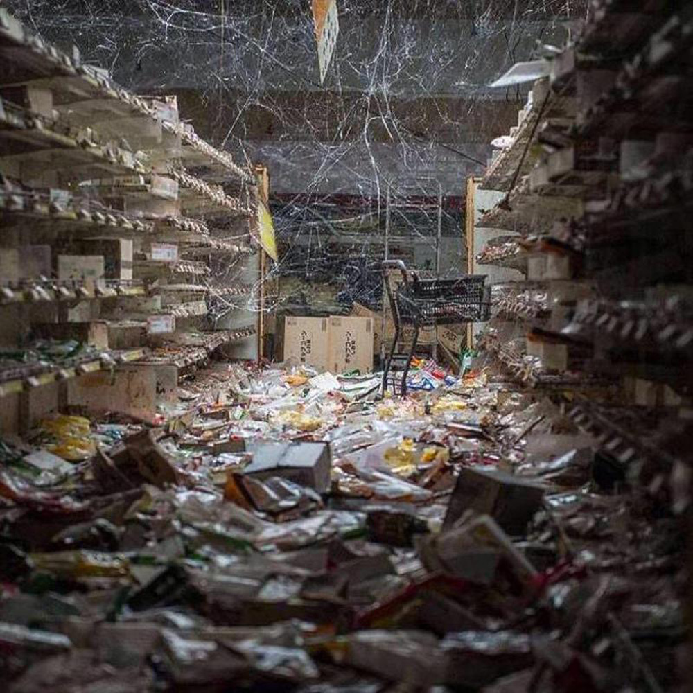 A dimly lit store aisle is filled with scattered debris and empty shelves. The floor is covered with various packages, and cobwebs hang from the ceiling, giving an abandoned appearance.