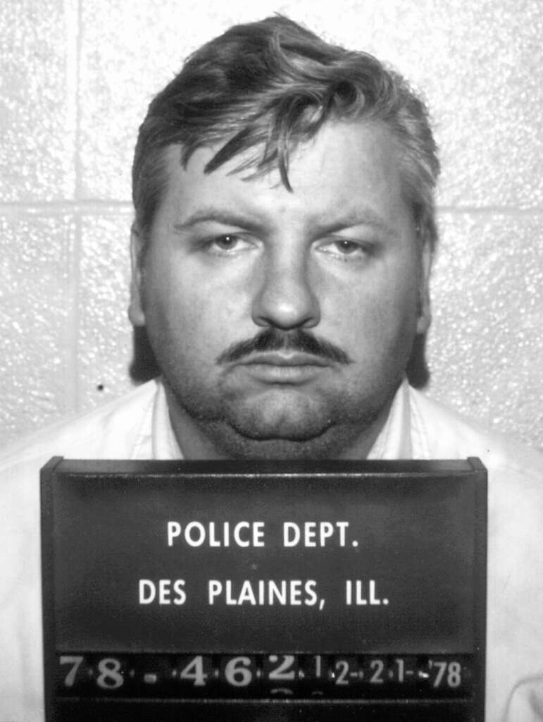 A black and white mugshot of an unidentified man with a mustache, wearing a collared shirt. He stares directly at the camera. The sign reads "Police Dept. Des Plaines, Ill." with numbers and a date "2-21-78" below.