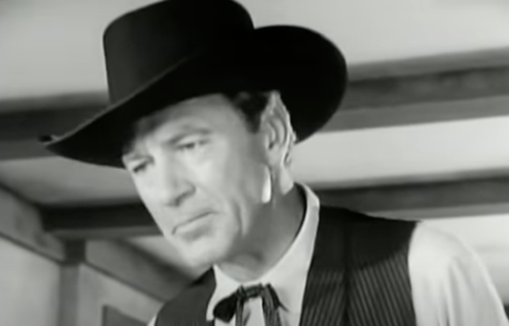 A black-and-white image of a man in a cowboy hat, vest, and tie looking down, with a neutral expression. The setting appears indoors, with wooden beams visible in the background.