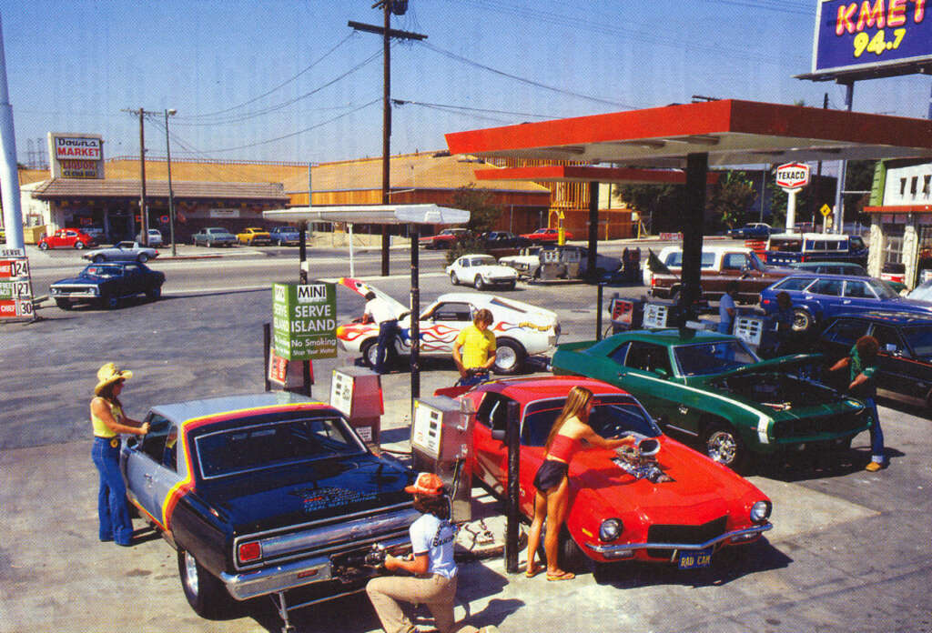 Retro scene at a gas station with vintage cars being refueled. People in casual 1970s attire, including a man with a cowboy hat and a woman in a red bikini, are standing by their colorful, classic cars. A market and buildings are in the background.