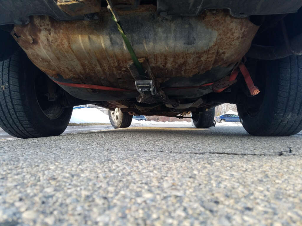 A low-angle view of the underside of a vehicle parked on asphalt, showing a rusty exhaust system and a metal component secured with straps. The tires are visible, and there are trees and snow in the background.