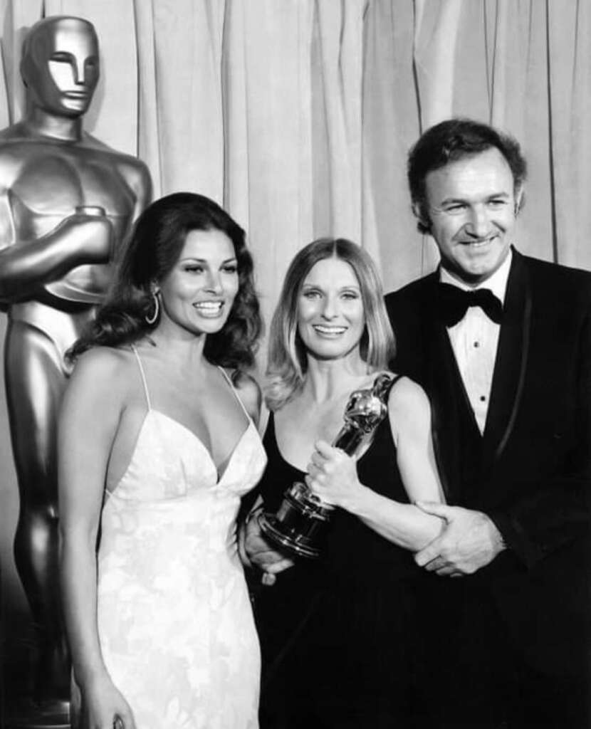 Three people in formal attire pose with an Oscar statue. One person in the center holds an Oscar award, flanked by a woman smiling in a white dress and a man in a tuxedo. They stand in front of a curtain backdrop next to a large Oscar statue.