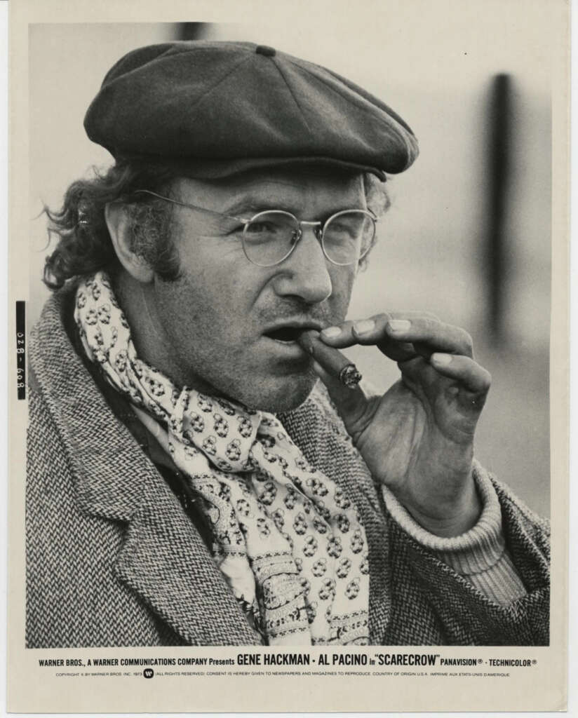 A man in glasses and a cap holds a cigar to his mouth. He's wearing a patterned scarf and a jacket. The image is black and white, with text at the bottom detailing cast and production information for the film "Scarecrow.