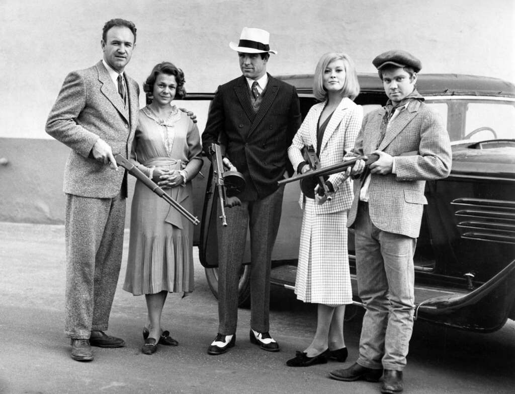 Five people dressed in 1930s-style clothing stand in front of a vintage car, holding period-appropriate weapons. The man in the center wears a suit and fedora. The group poses confidently for the camera.