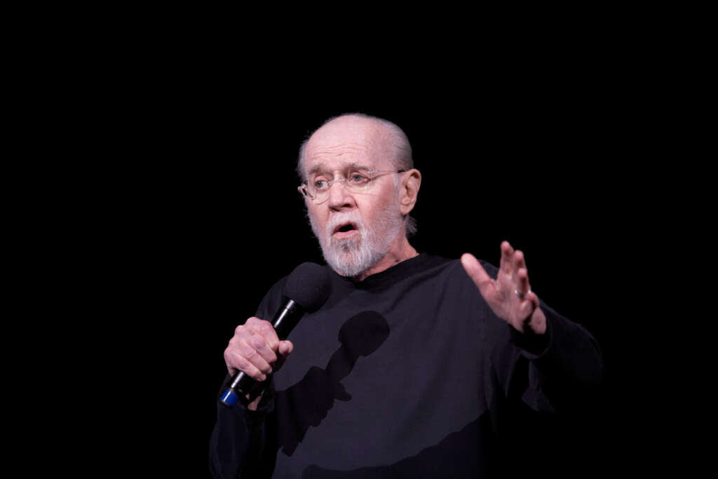 An elderly man with a goatee and glasses stands against a black background, holding a microphone. He's wearing a black shirt and has an expressive look, with one hand gesturing outward.