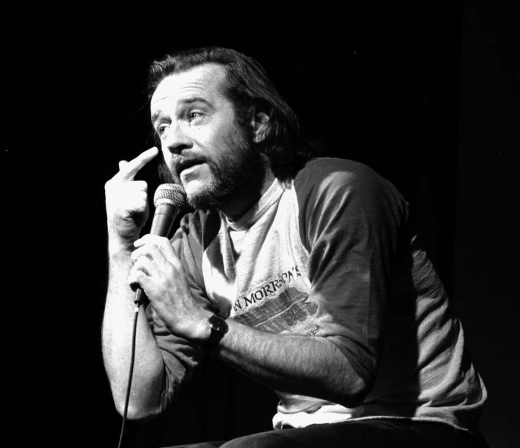 Comedian on stage holding a microphone, wearing a casual long-sleeve shirt. He gestures with his left hand near his head, engaging with the audience. The background is dark, focusing the light on him.