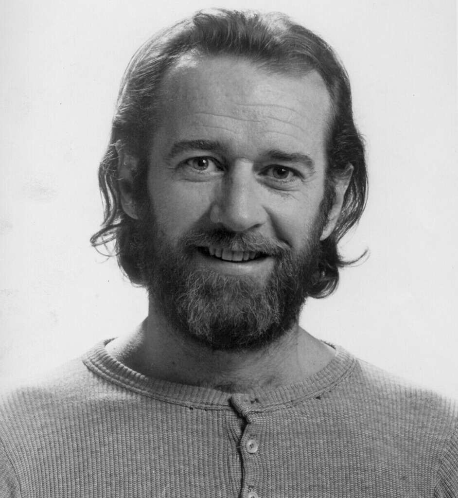 A black-and-white photo of a man with shoulder-length hair and a beard, smiling warmly. He is wearing a buttoned thermal shirt, and the background is plain.