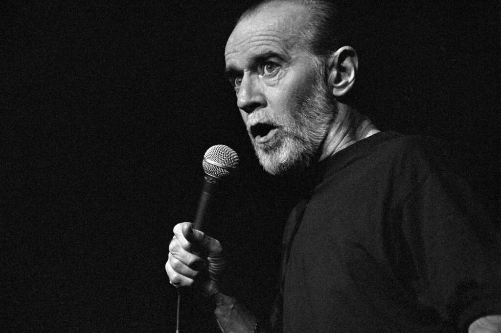 A person with a beard is holding a microphone, appearing to speak or perform on stage. The image is in black and white, emphasizing the dramatic lighting and expression.