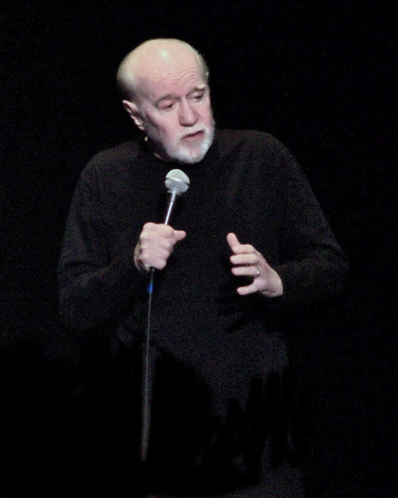 Elderly man with a bald head and white beard holds a microphone, wearing a black outfit, standing against a dark background, appearing to speak or perform.