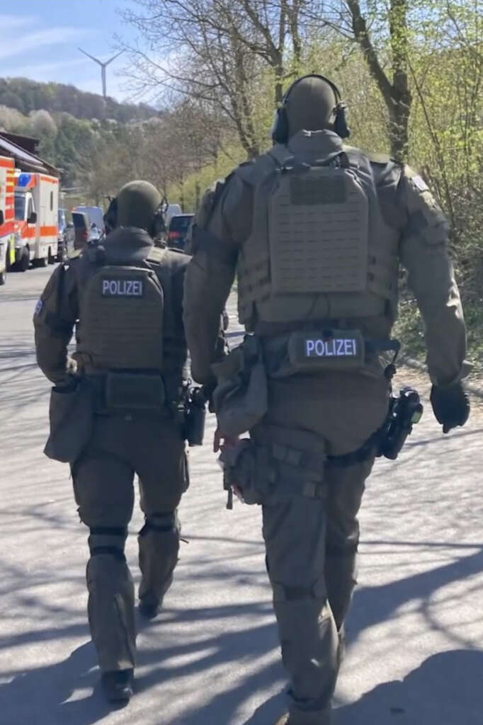 Two heavily armored police officers walk on a sunlit road in tactical gear labeled "Polizei." Trees and vehicles, including an emergency vehicle, are visible in the background. A wind turbine can be seen on a distant hill.