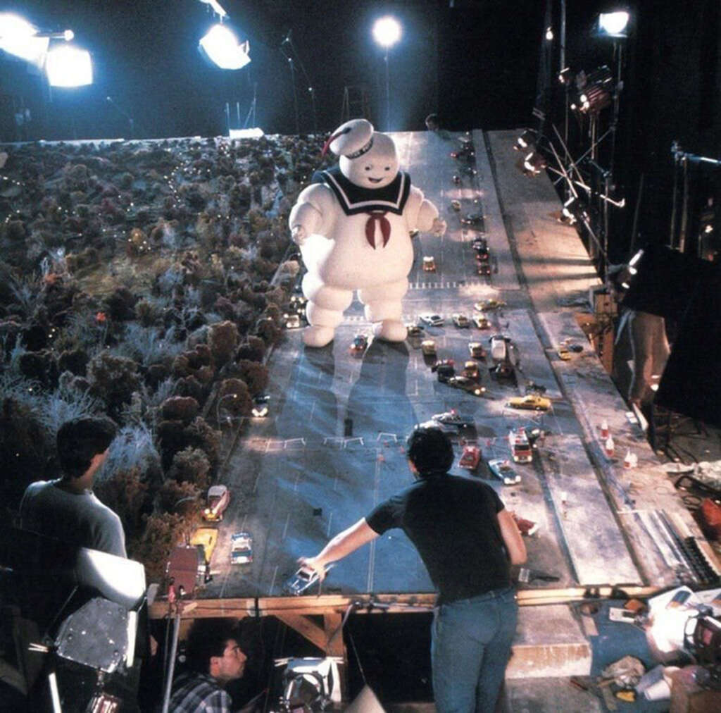 Behind-the-scenes view of a movie set with a large Stay Puft Marshmallow Man model towering over a miniature cityscape. Crew members adjust the scene, surrounded by cameras and lighting equipment.