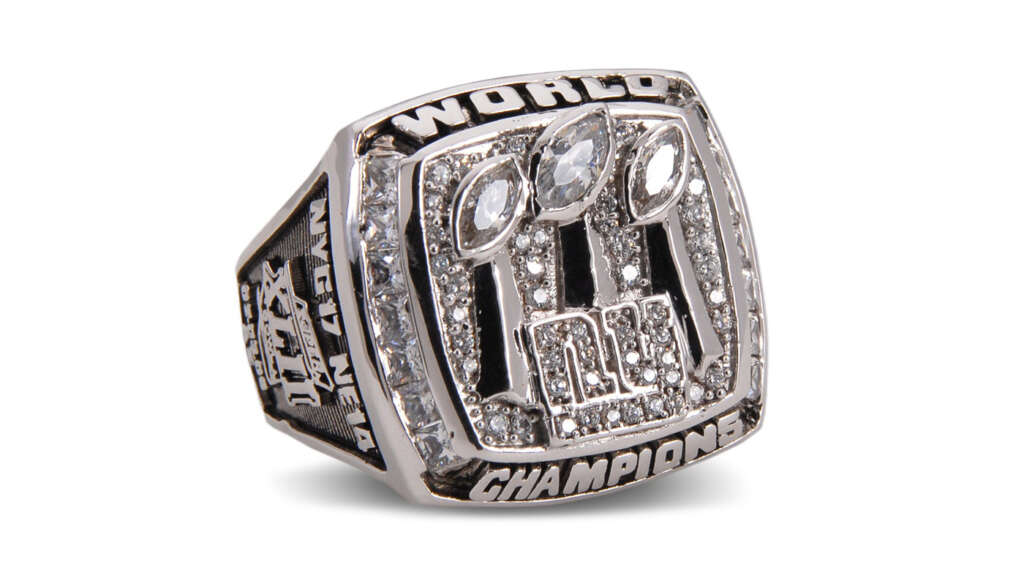 A silver championship ring adorned with three large football-shaped gemstones set above an NFL logo. The words "World Champions" are engraved on the top and bottom, with additional text on the sides. The ring is shiny and elaborately decorated.