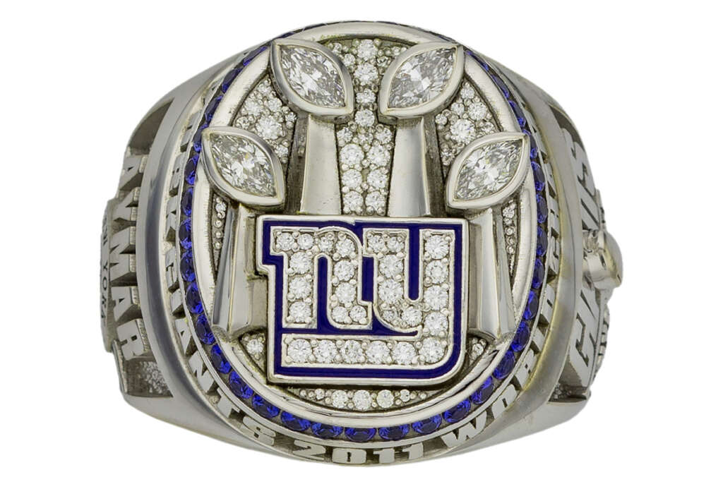 A New York Giants championship ring featuring the team's logo in blue and white, surrounded by diamond accents. The top has three football-shaped diamonds. Engravings and additional diamonds adorn the silver band.