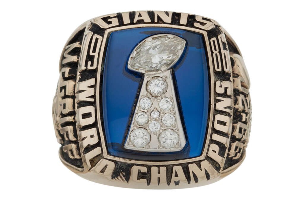 A championship ring featuring the word "Giants" at the top. The center displays a trophy design with multiple diamonds on a blue background. The words "World Champions" surround the trophy, with years visible on the sides.