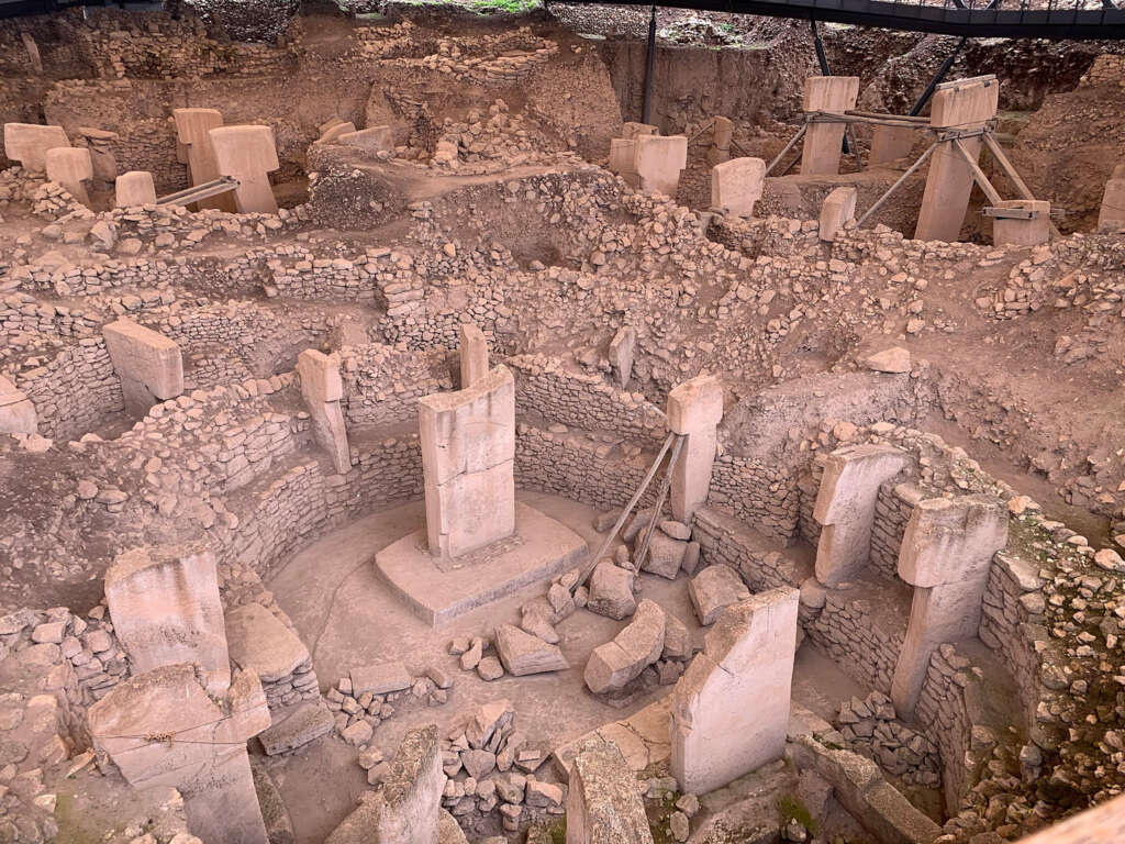 Ancient archaeological site featuring large stone pillars arranged in circular formations. The stones are intricately placed and some are supported by wooden beams. The site is partially excavated with surrounding dirt and rock debris.