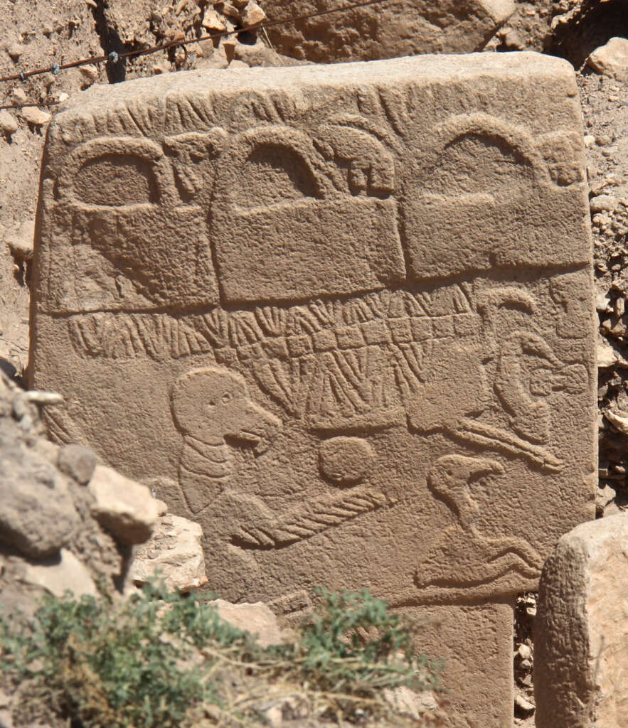 A weathered stone slab with ancient carvings, featuring abstract shapes and symbols, possibly depicting animals and other figures. The slab is surrounded by rocks and sparse greenery in an outdoor archaeological setting.