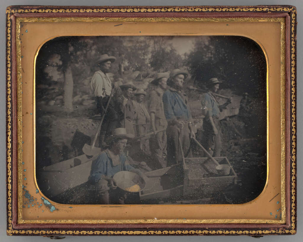 A vintage photograph shows a group of men wearing hats and work clothes standing by a riverbank. Some hold shovels and pans, suggesting they are gold prospectors. Trees are visible in the background, and the scene is framed with an ornate border.