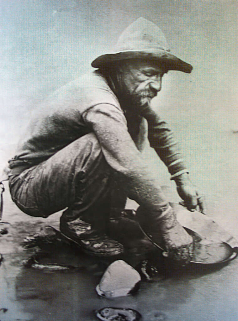 A black-and-white photo of an elderly man squatting by a river, panning for gold. He wears a wide-brimmed hat and rugged clothing, focused on his task with his hands submerged in the water, surrounded by stones.