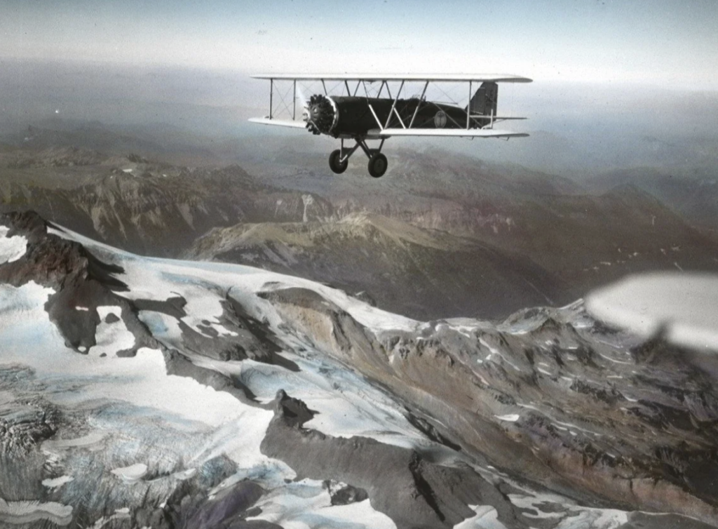 A vintage biplane flies over a mountainous landscape with snow-covered peaks and rugged terrain. The sky is clear, creating a sense of openness and freedom.