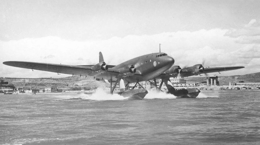 A vintage four-engine seaplane with pontoons makes a water landing. The aircraft, set against a backdrop of hills and buildings, has a sleek fuselage and is creating splashes in the water. The sky is partly cloudy.