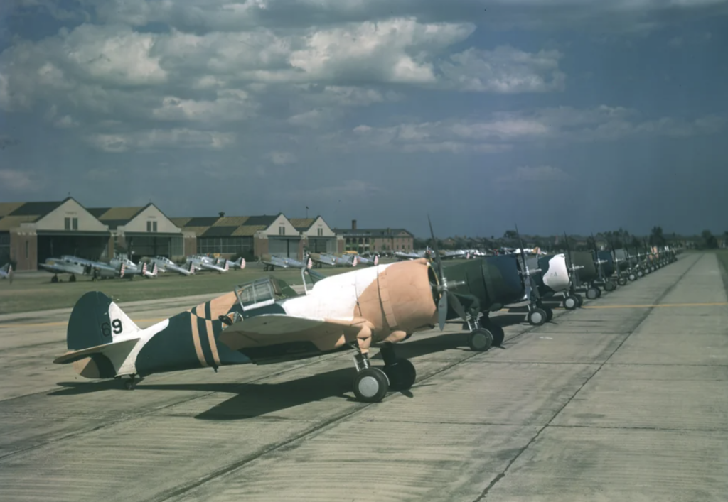 A line of vintage military aircraft parked on a runway, with several hangars in the background under a partially cloudy sky. The planes are painted in various camouflage patterns, and more planes are visible in the distance.