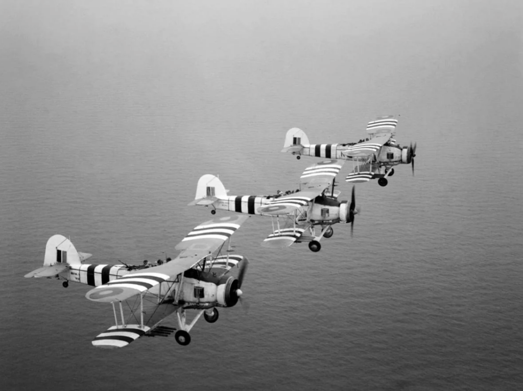 Four vintage biplanes in flight over a body of water. The aircraft feature distinct striped patterns on their wings and fuselages. The view is from a side angle, capturing their formation in an overcast sky.