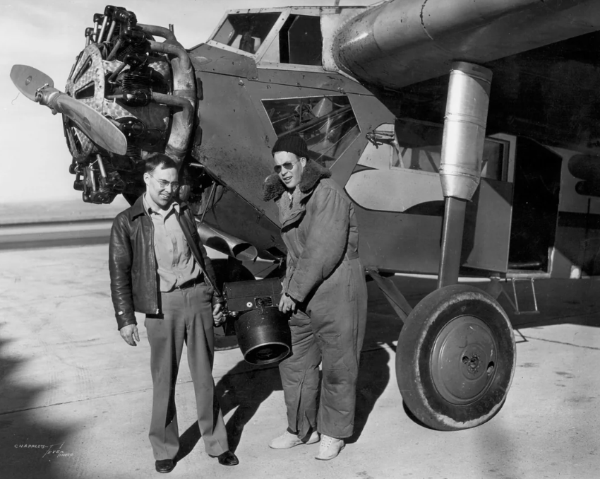 Two men stand next to an airplane's engine. One wears a leather jacket and the other is in a flight suit and goggles, holding a large camera. The plane's wing and propeller are visible. The setting appears to be an airfield.