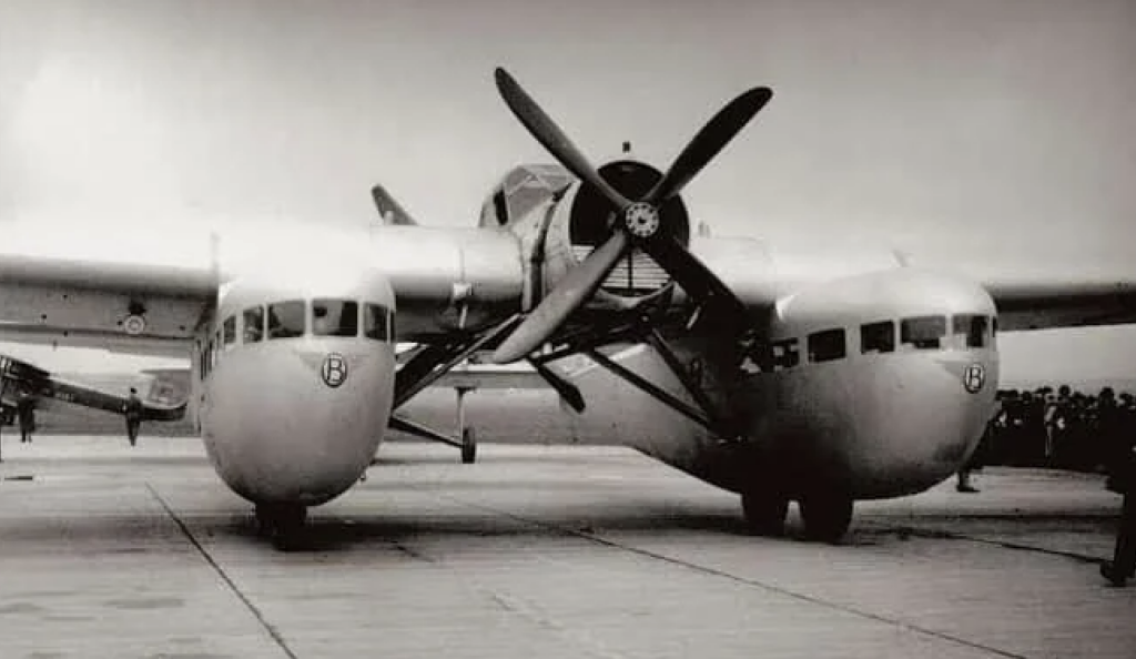 Black and white photo of a vintage twin-fuselage airplane on a tarmac. The aircraft has a large central propeller and round windows on each fuselage. People are gathered nearby, with an overcast sky in the background.