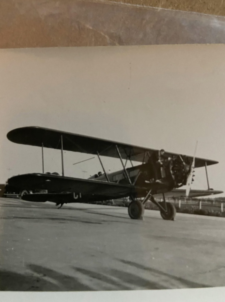 A vintage black and white photo of a biplane parked on a runway. The plane has two wings stacked vertically and a visible propeller. A person is seated in the cockpit. The background shows trees and a clear sky.