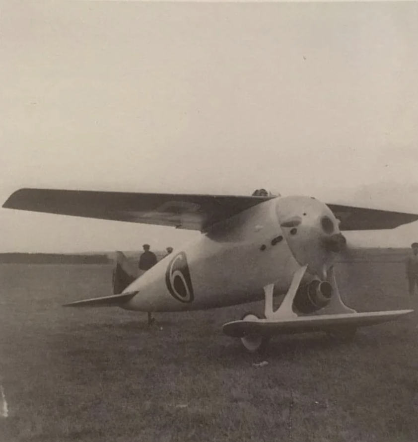 A vintage black and white photo of a single-engine monoplane with large wings, parked on a grassy field. Three people stand nearby, observing the aircraft. The tail features a distinctive circular symbol.