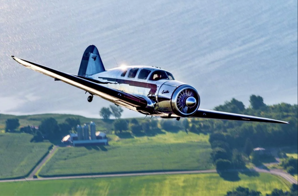 A vintage airplane with a polished metal exterior flies over a lush green landscape and a body of water. The plane's propeller is spinning, and the sun glints off its shiny surface. Farmland and trees are visible below.