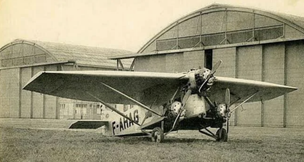 A vintage single-engine airplane with the registration "F-AJHG" is parked on grass before two large hangars. The aircraft features a high wing and open cockpit design, typical of early aviation models.