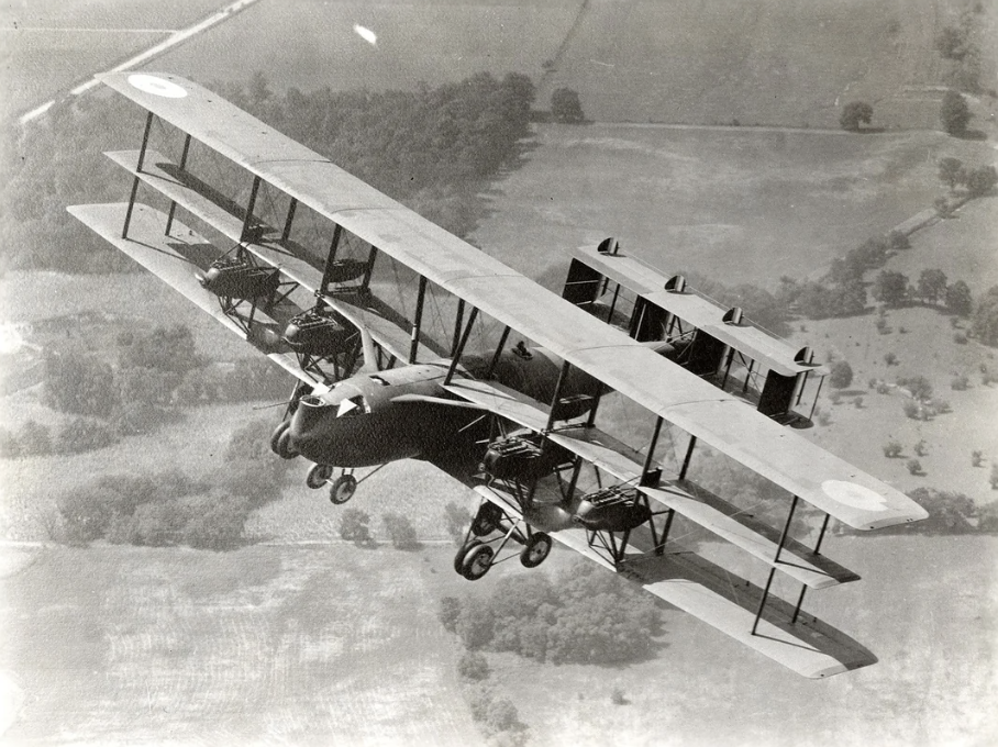 A vintage black and white photograph of a large triplane aircraft flying over a landscape. The aircraft has multiple wings and open cockpits, with fields and trees visible below.