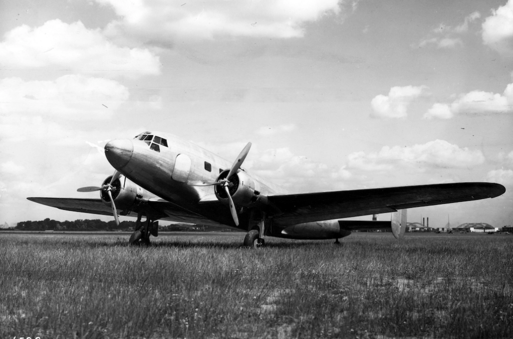 A vintage twin-engine propeller airplane is parked on a grassy field under a partly cloudy sky. The aircraft features a sleek fuselage with a cockpit, angled wings, and two propellers. Industrial buildings are visible in the distant background.