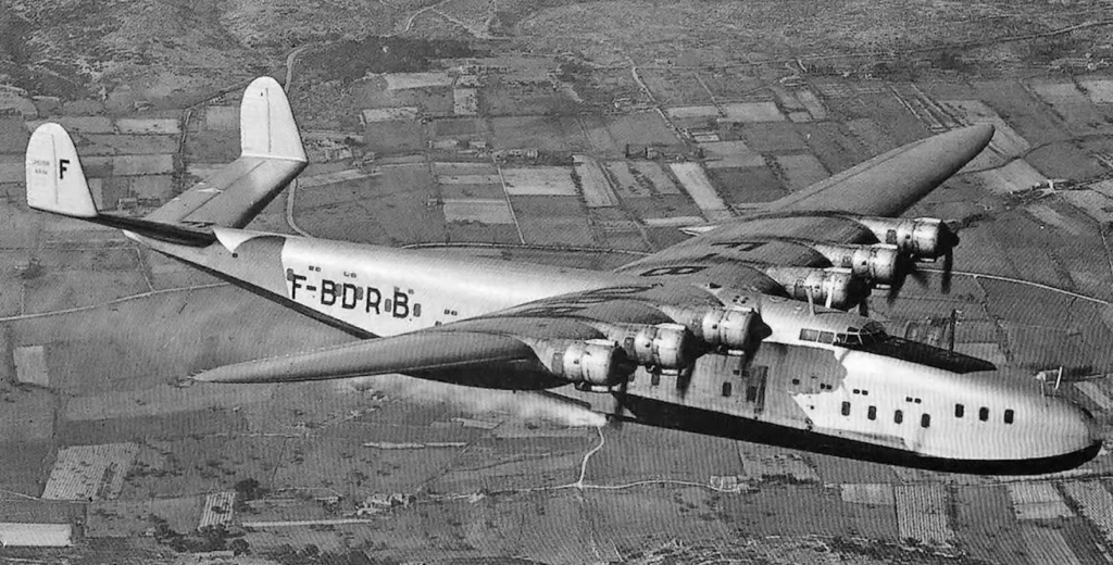 A large, vintage seaplane with four engines flying over a patchwork landscape. The aircraft has a silver body with the registration "F-BDRB" visible on the side. The terrain below features fields divided by hedges and roads.