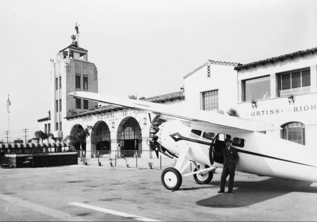 A black-and-white photo depicting a small vintage airplane parked on a tarmac near a control tower labeled "Grand Central Air Terminal." A man stands by the plane's wing, with buildings in the background.