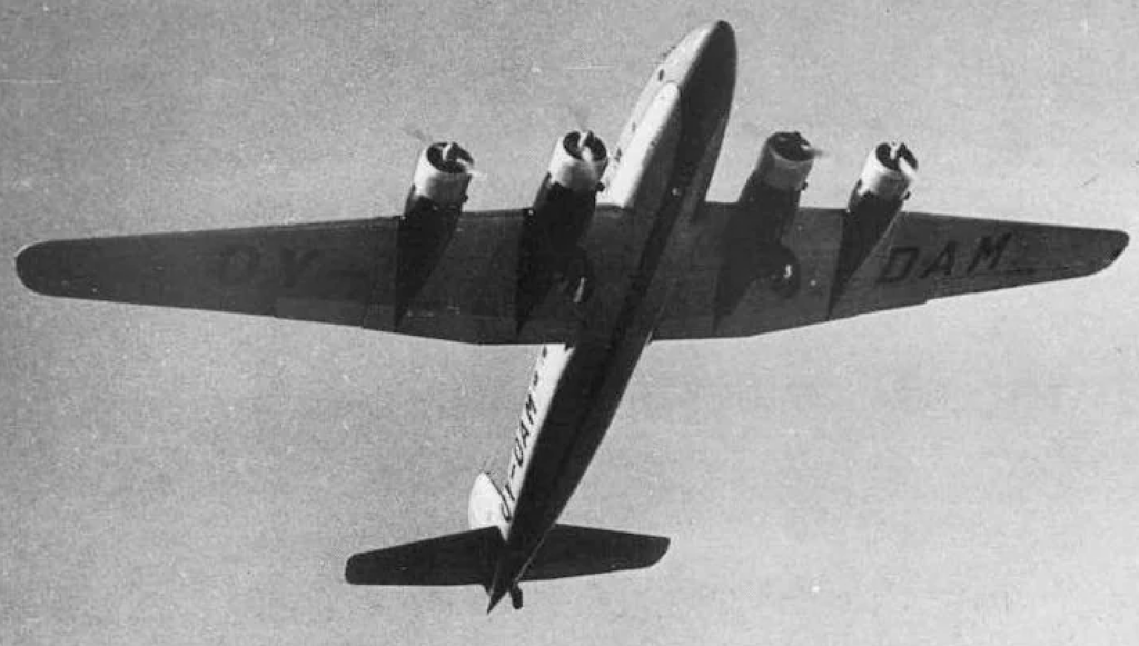 A black and white photo of a four-engine propeller aircraft flying overhead. The plane has letters on the underside of its wings and appears to be in mid-flight in a clear sky.