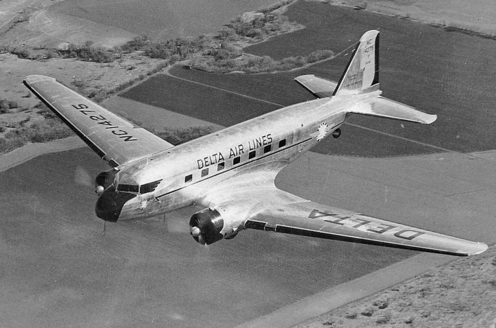 Historic black and white image of a Delta Air Lines plane in flight. The aircraft is a propeller-driven model from the mid-20th century, flying over a patchwork landscape of fields. The registration number NC14715 is visible on the wing and tail.