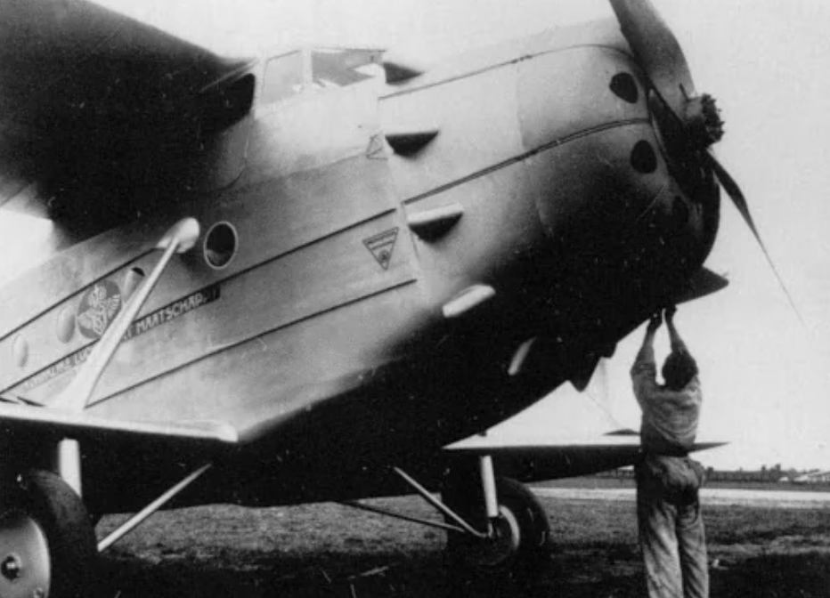 A person stands next to the large front engine of a vintage biplane, working on or inspecting it. The plane has a robust, classic design, and is on grassy ground, highlighting its metallic structure and details. Black and white photo.