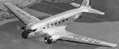 Black and white photo of a vintage Delta Airlines propeller plane in flight over farmland. The aircraft has a sleek, reflective surface with the airline's name visible on the side and wing. Trees and fields are seen below.