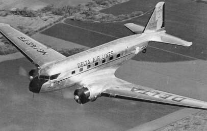 Black and white photo of a vintage Delta Airlines propeller plane in flight over farmland. The aircraft has a sleek, reflective surface with the airline's name visible on the side and wing. Trees and fields are seen below.