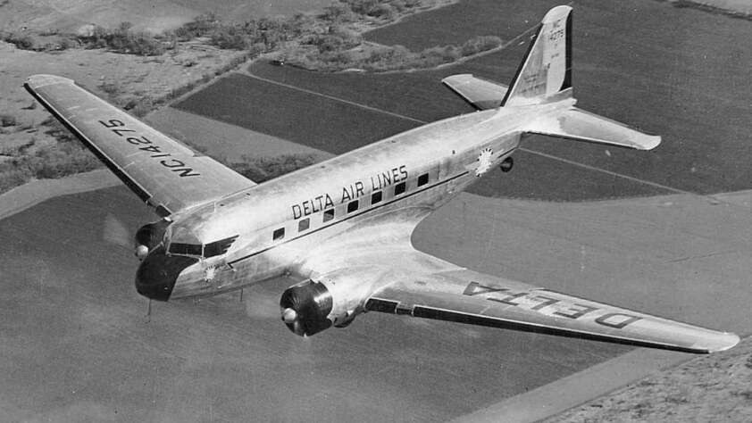 Black and white photo of a vintage Delta Airlines propeller plane in flight over farmland. The aircraft has a sleek, reflective surface with the airline's name visible on the side and wing. Trees and fields are seen below.