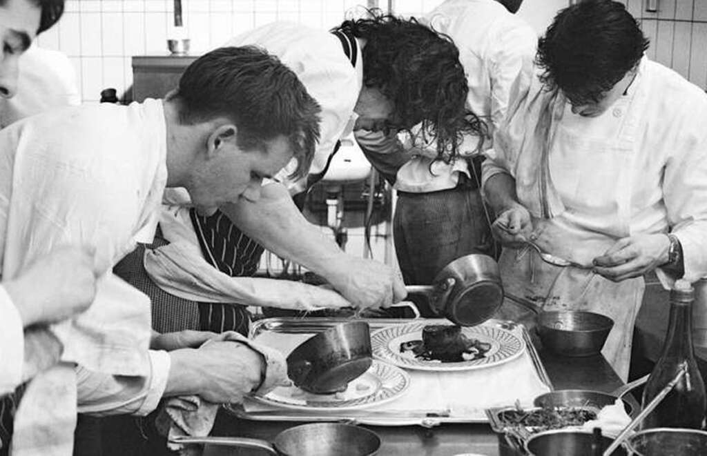 Black and white photo of chefs in a kitchen, focused on plating dishes. They are pouring sauce and arranging ingredients with precision, surrounded by kitchen tools and ingredients. The scene conveys intense concentration and teamwork.