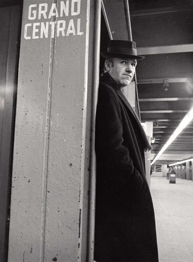 A man wearing a dark coat and hat leans against a pillar marked "Grand Central" in a dimly lit corridor. He gazes thoughtfully into the distance, with an empty walkway extending behind him.