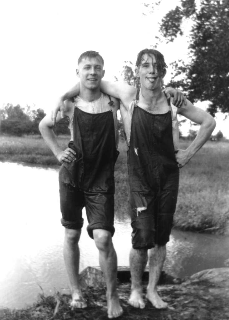 Two men in wet overalls stand barefoot on a rock by a river, smiling with one draping an arm around the other. Both are soaked, indicating they have been in the water. Trees and grass are visible in the background.