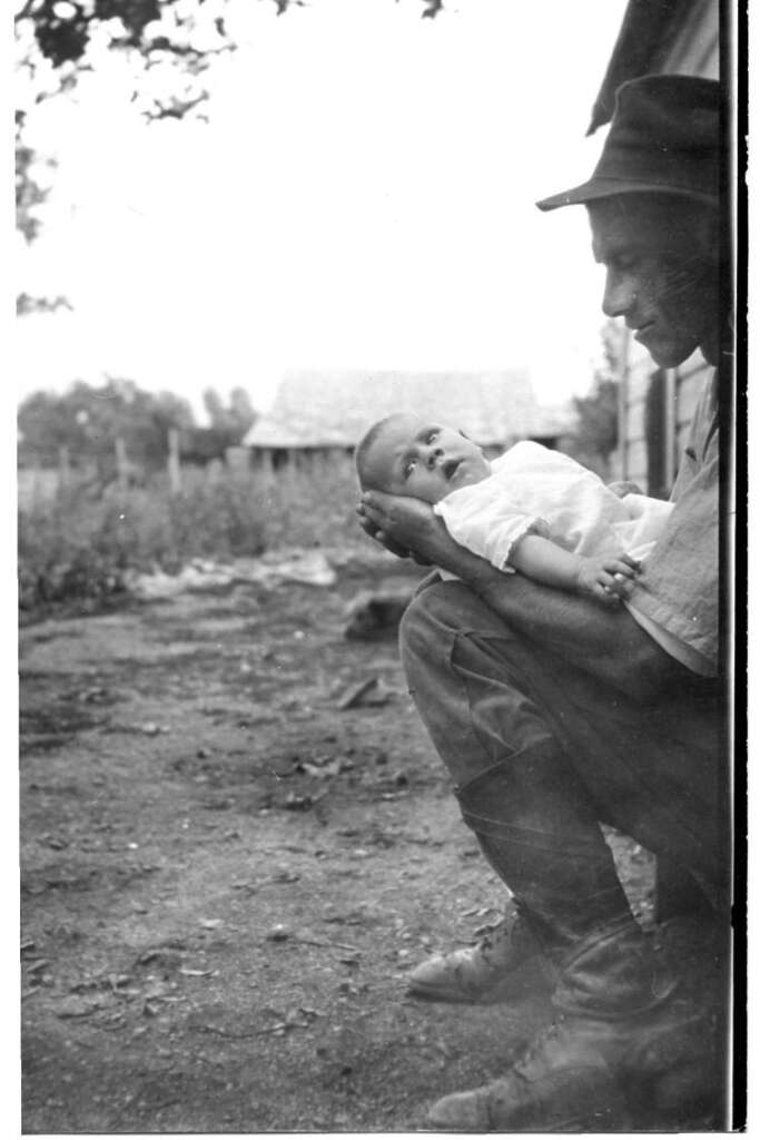 A man wearing a hat crouches beside a building, cradling a baby in his arms. The baby looks up at him. In the background, there is a rural landscape with a house and trees.