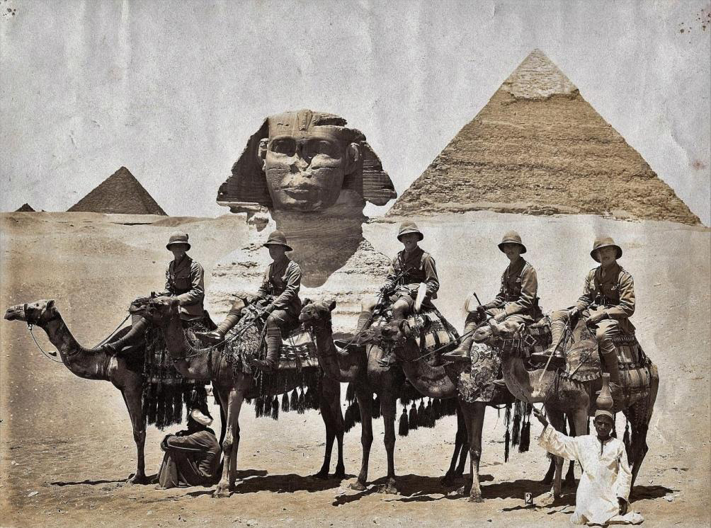 A vintage photograph shows a group of men in early 20th-century attire riding camels in front of the Sphinx and pyramids in Egypt. A local guide stands beside them, while one person tends to the camels. The scene conveys a sense of historical exploration.