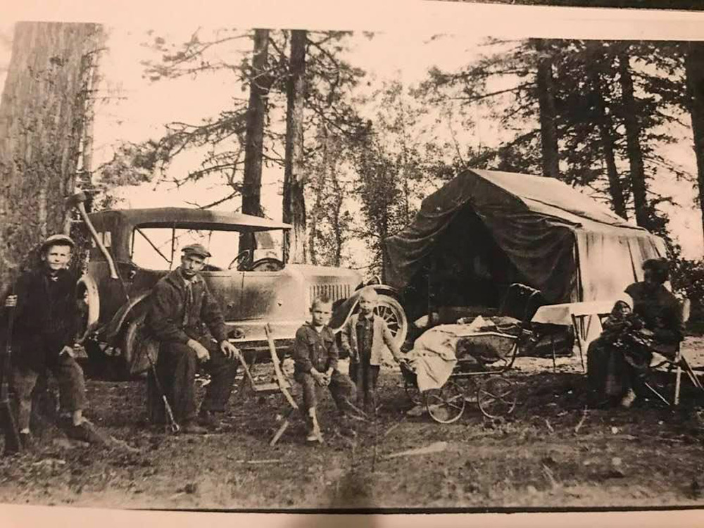 A vintage black and white photo of a family camping in a forest. They are sitting and standing around a classic car and a canvas tent. A baby carriage and makeshift table are nearby among the tall trees.