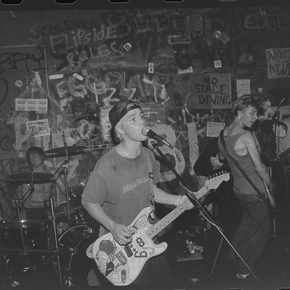 A black and white photo of a band performing live in a graffiti-covered venue. The guitarist sings into a microphone, wearing a T-shirt and cap. The drummer plays in the background, and another musician is on the right, playing guitar.